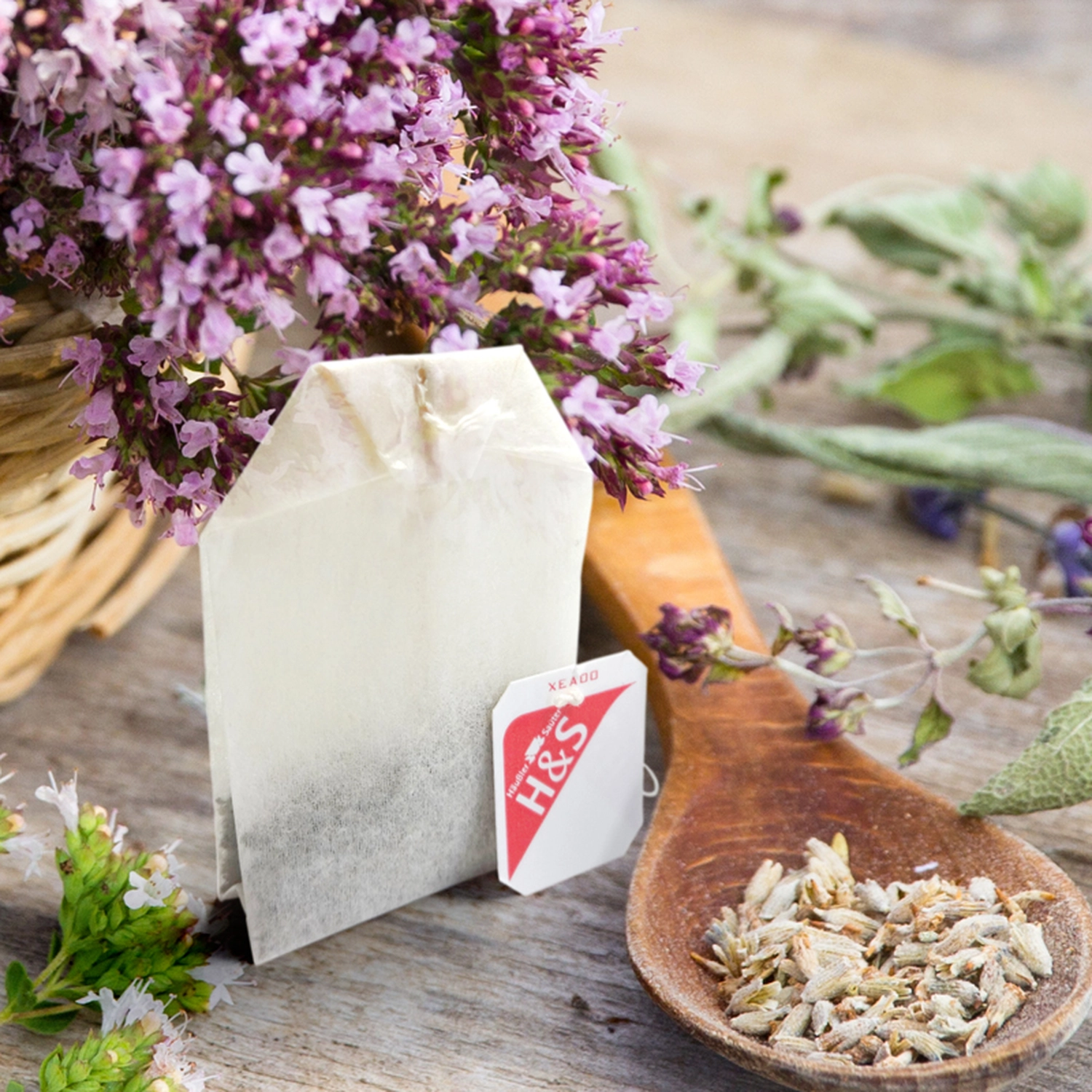 Linden-Apotheke - Pharmazeutische Dienstleistungen & Online-Bestellung - Ein weißer Teebeutel aus Papier mit einem rot-weißen H&S-Etikett liegt neben einem Holzlöffel, der mit losen getrockneten Kräutern und frischen lila Blüten gefüllt ist.